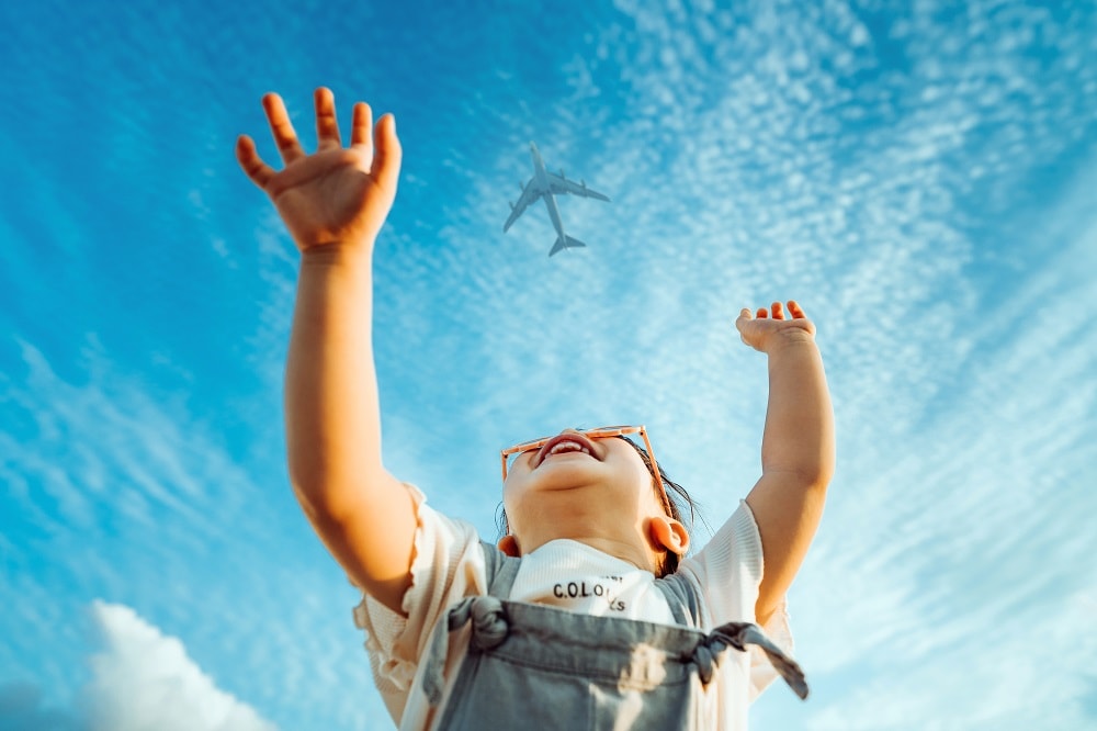 Happy little Asian girl with flower-shaped sunglasses smiling joyfully and raised her hands waving to the aeroplane in the clear blue sky Getty Images
