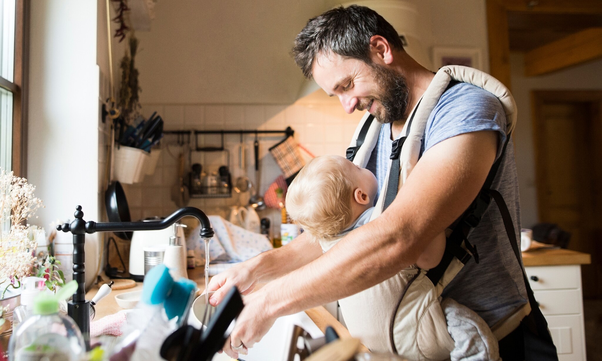 vader draagt baby in draagzak of draagdoek terwijl hij in de keuken bezig is Getty Images