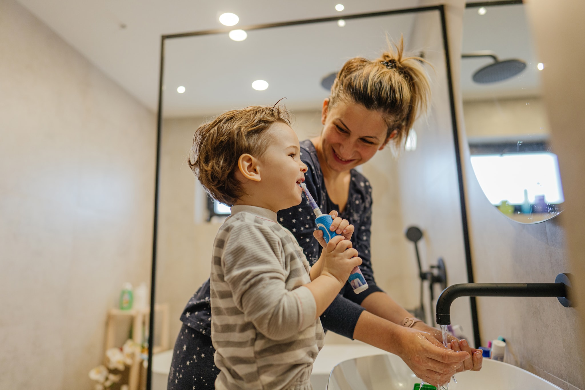 Photo of a little boy brushing his teeth in the bathroom after waking up. His mother is right beside him, giving him good example.  Frozen elektrische tandenborstel van Oral-B - Disney ©