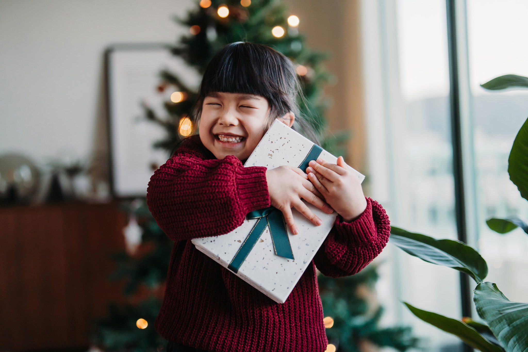 Happy little Asian girl hugging a box of Christmas present on Christmas morning, showing her excitement. Standing in front of Christmas tree in the living room at home. Christmas lifestyle theme. Celebrating Christmas. Holiday and festive vibes Getty Images