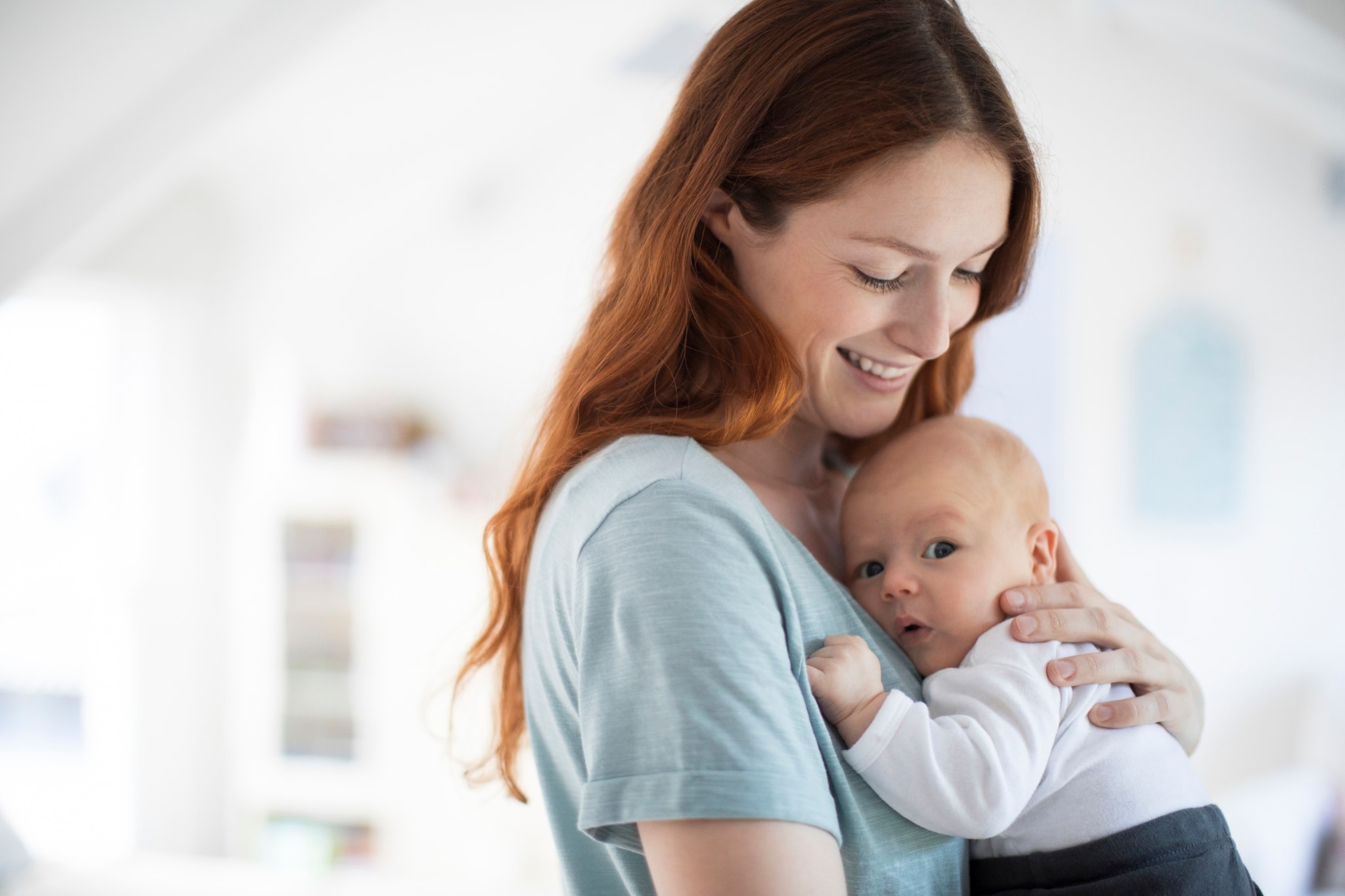 Beautiful mother carrying newborn baby boy in bedroom. Happy redhead woman is standing with cute son. They are at home. Getty Images